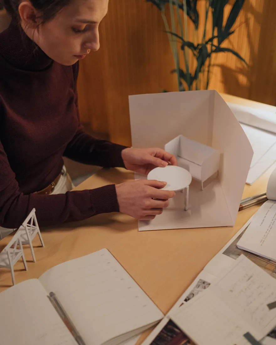 Mesa de trabajo con recortes de cartón, planos y piezas de maqueta en preparación bajo luz lateral